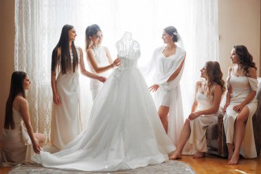 A group of women are posing in front of a wedding dress. The dress is white and has a long train. The women are dressed in white and are sitting on a couch. Scene is happy and celebratory