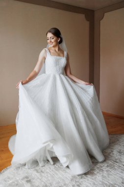 Beyaz elbiseli bir gelin resim için poz veriyor. O bir peçe takıyor ve kollarını açıyor. Resmin mutlu ve kutlama havası var, gelinin hayatındaki özel bir anı yakalıyor.