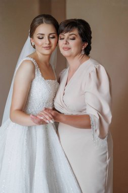 Bir gelin ve annesi birlikte duruyorlar. Gelin beyaz bir elbise giyiyor ve anne pembe bir elbise giyiyor. Sahne mutlu ve kutlama dolu.