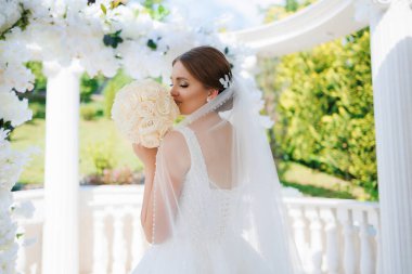 Bir gelin, elinde bir buket çiçek olan bir resim için poz veriyor. Beyaz bir elbise ve duvak takıyor. Sahne arka planda beyaz kemerli bir bahçede geçiyor.