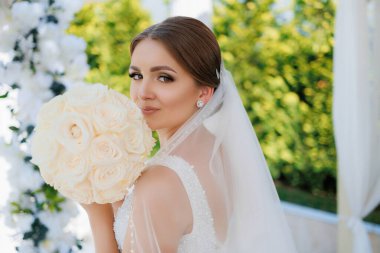 Bir gelin bir buket beyaz gülle fotoğraf çektirmek için poz veriyor. Beyaz bir elbise ve duvak takıyor. Gelin geleneksel bir düğün kıyafeti giydiği için, zarafet ve romantizm kavramı