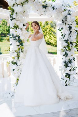 Beyaz bir kemerin önünde elinde çiçeklerle bir gelin duruyor. Beyaz bir elbise ve duvak takıyor.