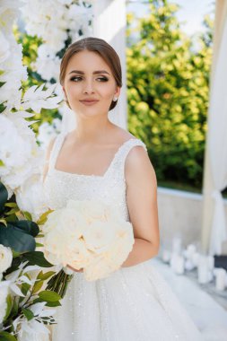 Bir kadın elinde bir buket beyaz çiçekle beyaz bir kemer yolunun önünde duruyor. Beyaz bir elbise giyiyor ve saçını arkaya çekiyor.