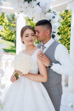 Bir gelin ve damat beyaz bir kemerin önünde poz veriyorlar. Gelin bir buket çiçek tutuyor ve damat yelek giyiyor. Sahne mutlu ve romantik.