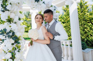 Bir gelin ve damat beyaz bir kemerin altında duruyor. Gelin bir buket çiçek tutuyor ve damat yelek giyiyor. Sahne bir bahçede ya da parkta geçiyor, yakınlarda ağaçlar ve bir bank var.