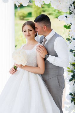 Bir gelin ve damat beyaz bir kemerin önünde poz veriyorlar. Gelin bir buket tutuyor ve damat kravat takıyor. Sahne mutlu ve romantik.