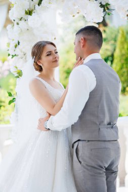 Bir gelin ve damat bir düğün töreninde birbirlerini kucaklıyorlar. Gelin beyaz bir elbise giyiyor ve damat gri bir yelek giyiyor. Sahne aşkla dolu.