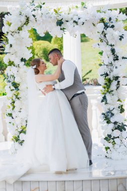 Bir gelin ve damat beyaz bir kemerin altında öpüşüyor. Gelin beyaz bir elbise giyiyor ve damat gri bir yelek giyiyor.