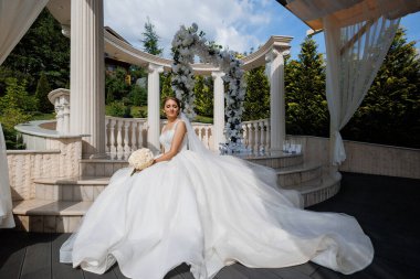 Beyaz elbiseli bir kadın, beyaz bir kemerin önündeki taş bir merdivende oturuyor. Elinde bir buket çiçek tutuyor.