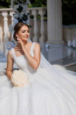 Gelinlik giymiş bir kadın elinde buketle mermer zeminde oturuyor. Gelin güzel beyaz bir elbise giydiği ve çiçeklerle çevrili olduğu için, zarafet ve kutlama kavramı