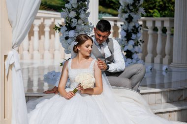 Gelin ve damat taş bir merdivende oturuyor, gelin elinde buket tutuyor. Sahne romantik ve samimi.