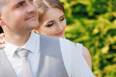 Bir gelin ve damat yemyeşil bir alanda birbirlerine sarılıyorlar. Gelin beyaz bir elbise giyiyor ve damat gri bir takım elbise giyiyor. Sahne aşk ve mutluluktur.