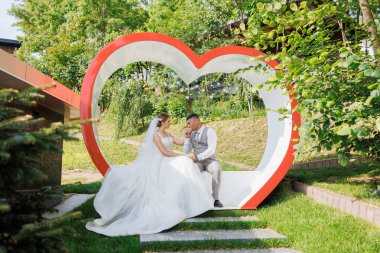 Bir gelin ve damat parktaki kırmızı kalp şeklinde bir bankta oturuyorlar. Romantik ve samimi bir sahne. Çiftler birlikte eğleniyor.