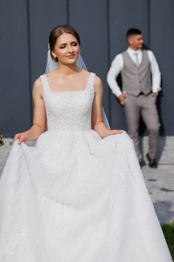 Bir gelin ve damat binanın önünde fotoğraf için poz veriyorlar. Gelin inci başlıklı ve duvağı olan beyaz bir elbise giyiyor. Damat gri bir yelek ve kravat takıyor.