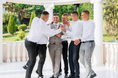 Bir grup adam birlikte duruyor, içlerinden biri kravat takıyor. Hepsi gülümsüyor ve birbirlerine eşlik etmekten zevk alıyor gibi görünüyorlar.