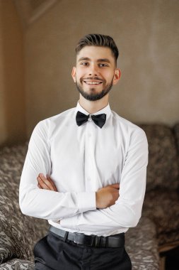 Beyaz gömlekli ve siyah papyonlu bir adam gülümsüyor ve kollarını kavuşturmuş duruyor. Resmin neşeli ve dost canlısı bir havası var. Adamın fotoğrafa poz vermesi gibi.