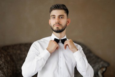 Beyaz gömlekli ve siyah papyonlu bir adam dışarı çıkmaya hazırlanıyor. Kahverengi bir duvarın önünde duruyor.