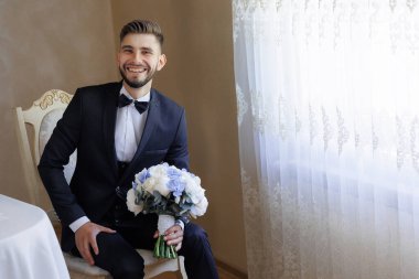 Takım elbiseli ve kravatlı bir adam kucağında bir buket çiçekle bir sandalyede oturuyor. Gülümsüyor ve mutlu.