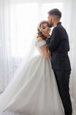 Bir gelin ve damat pencerenin önünde sarılıyorlar. Gelin beyaz bir elbise giyiyor ve damat siyah bir takım elbise giyiyor. Sahne mutlu ve romantik.