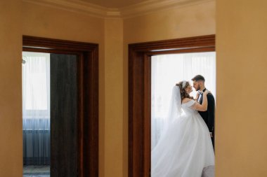 Gelin ve damat bir duvarın önünde duruyorlar. Gelin beyaz bir elbise ve damat siyah bir takım elbise giyiyor.