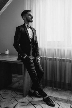 Takım elbiseli ve kravatlı bir adam pencerenin önünde duruyor. Papyon takıyor ve yüzünde ciddi bir ifade var.