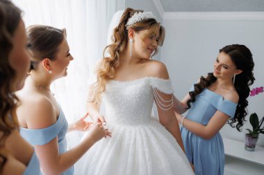 Bir gelin nedimeleriyle düğününe hazırlanıyor. Gelin, başında taç olan beyaz bir elbise giyiyor.