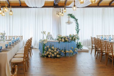 Mavi masalı ve beyaz perdeli büyük bir oda. Oda çiçeklerle süslenmiş ve üzerinde beyaz çiçekler olan mavi bir masası var. Masanın etrafında bir sürü sandalye var.