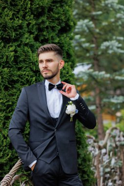 Siyah takım elbiseli ve beyaz gömlekli bir adam yeşil bir çalılığın önünde duruyor. Siyah bir papyon takıyor ve elinde bir çiçek tutuyor. Zarafet ve incelik kavramı