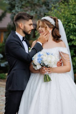 Bir gelin ve damat fotoğraf için poz veriyor. Gelin bir buket çiçek tutuyor ve damat onun yüzüne dokunuyor. Sahne mutlu ve romantik.