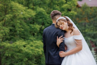 Bir gelin ve damat yemyeşil bir alanda birbirlerine sarılıyorlar. Damat takım elbise giyerken gelin beyaz bir elbise ve peçe takıyor. Sahne huzurlu ve romantik.