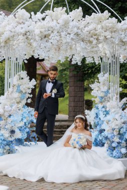Bir gelin ve damat ellerinde çiçeklerle beyaz bir kemer yolunun önünde poz veriyorlar. Gelin beyaz bir elbise giyiyor ve damat da takım elbise giyiyor.