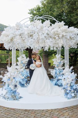 Bir gelin ve damat beyaz bir kemerin altında poz veriyorlar. Kemer çiçeklerle süslenmiş ve mavi-beyaz bir renk şemasına sahiptir. Çift resmi giyinmiş.