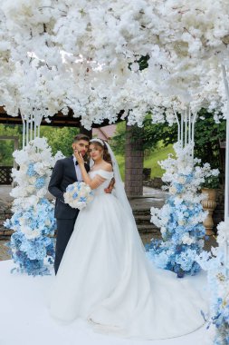 Bir gelin ve damat beyaz bir kemer yolunun önünde poz veriyorlar. Gelin beyaz bir elbise giyiyor ve damat bir takım elbise giyiyor. Kemer yolu beyaz çiçeklerle süslenmiş.