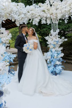 Bir gelin ve damat beyaz bir kemerin altında duruyor. Gelin beyaz bir elbise giyiyor ve damat bir takım elbise giyiyor. El ele tutuşup kameraya bakıyorlar.