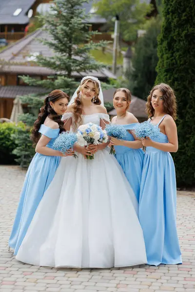 Bir gelin ve nedimeleri binanın önünde fotoğraf çektirmek için poz veriyorlar. Gelin beyaz bir elbise ve mavi bir duvak giyiyor.