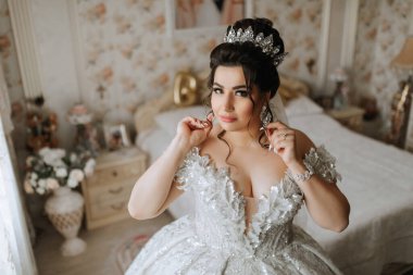 Kafasında taç olan beyaz elbiseli bir kadın. Küpe takıyor ve yatağın önünde duruyor.