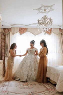 Üç kadın bir odada dikiliyor, biri gelinlik giyiyor. Diğer iki kadın düğün için hazırlanmasına yardım ediyor. Odanın bir sürü süsü var.