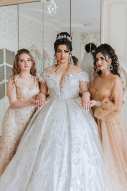 Üç kadın aynanın önünde poz veriyor. Kadınlardan biri kafasında taç olan beyaz bir elbise giyiyor. Diğer iki kadın altın elbiseler giyiyor.