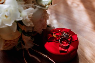 İçinde üç yüzük olan kırmızı bir kutu. Kutu ahşap bir masanın üzerine konur. Yüzükler altın ve elmas işlemeli. Arkadaki çiçekler beyaz.