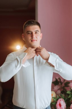 Beyaz gömlekli bir adam dışarı çıkmaya hazırlanıyor. Yakasını düzeltiyor ve kameraya bakıyor.