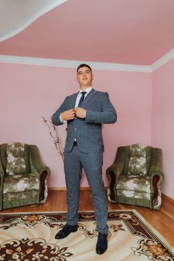 Takım elbiseli bir adam iki sandalyenin önünde duruyor. O bir kravat takıyor ve onu ayarlıyor. Odada pembe bir duvar ve yeşil bir sandalye var.