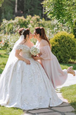 İki kadın çimlerin üzerinde oturuyor, biri gelinlik giyiyor. Diğer kadın elinde bir buket çiçek tutuyor. Sahne neşeli ve neşeli.