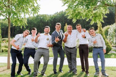 Bir grup adam parkta fotoğraf çektirmek için poz veriyor. Hepsi takım elbise ve kravat takmış ve içlerinden biri sarı papyon takıyor. Sahne neşeli ve neşeli.