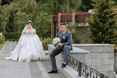 Bir gelin ve damat binanın önünde duruyor. Gelin beyaz bir elbise giyiyor ve damat bir takım elbise giyiyor. Gelin bir buket tutuyor ve damat da bir buket tutuyor.