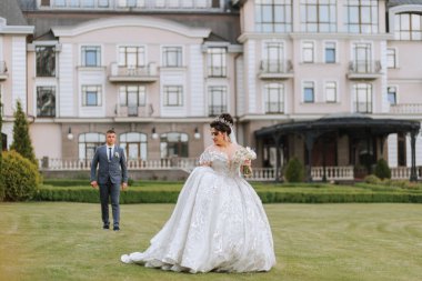 Bir gelin ve damat büyük bir binanın önünde çimenli bir yolda yürüyorlar. Gelin uzun beyaz bir elbise giyiyor ve damat bir takım elbise giyiyor. Sahne romantik ve resimli.