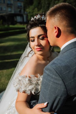 Bir gelin ve damat parkta birbirini kucaklıyor. Gelin beyaz bir elbise giyiyor ve damat bir takım elbise giyiyor. Sahne romantik ve mutlu.