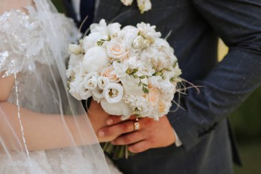 Bir gelin ve damat bir buket beyaz çiçek tutuyorlar. Gelin peçe takıyor ve damat takım elbise giyiyor. Sahne mutlu ve romantik.