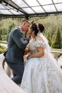 Gelin ve damat merdivenlerde öpüşürler. Gelin peçeli beyaz bir elbise giyiyor ve damat takım elbise giyiyor. Sahne romantik ve mutlu.