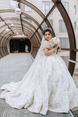 Beyaz elbiseli bir kadın elinde bir buket çiçek tutuyor. Bir tünelde ya da koridorda duruyor, muhtemelen bir binada. Sahne zarafet ve entelektüellik hissi veriyor.