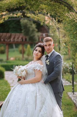 Gelin ve damat, çardağın önünde fotoğraf çektirmek için poz veriyorlar. Gelin beyaz bir elbise giyiyor ve damat takım elbise giyerken elinde bir buket tutuyor. Sahne mutlu ve romantik.